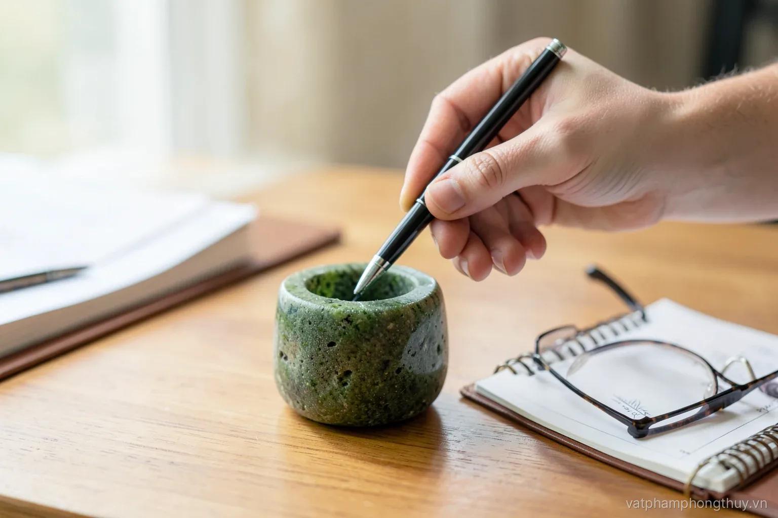 Close-up of a hand dropping a pen into a green stone pen holder on a tidy wooden desk beside a notebook and reading glasses, soft daylight