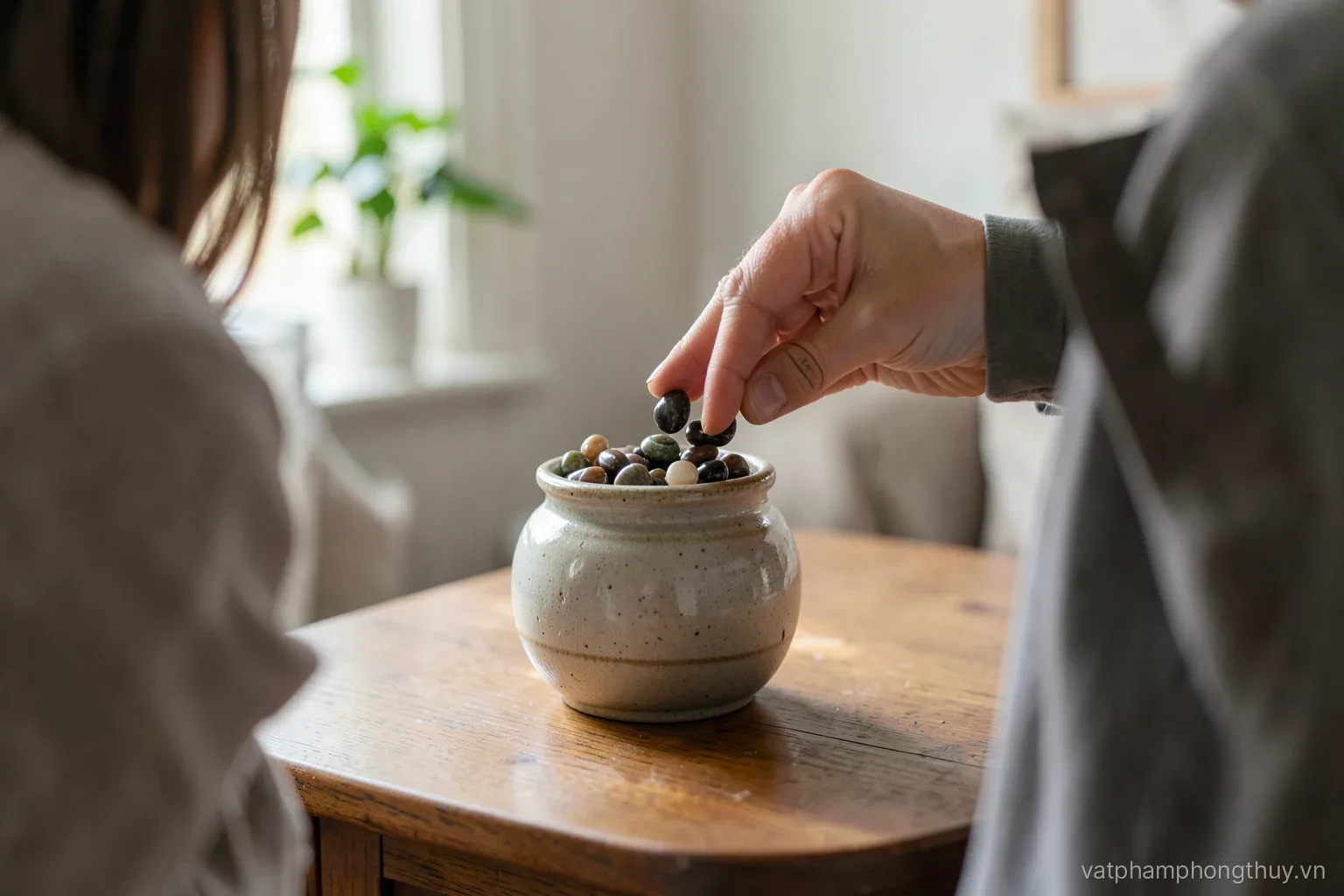 A person seen from behind pouring small polished stones into a round lidded ceramic jar on a wooden cabinet in a quiet living room, daylight