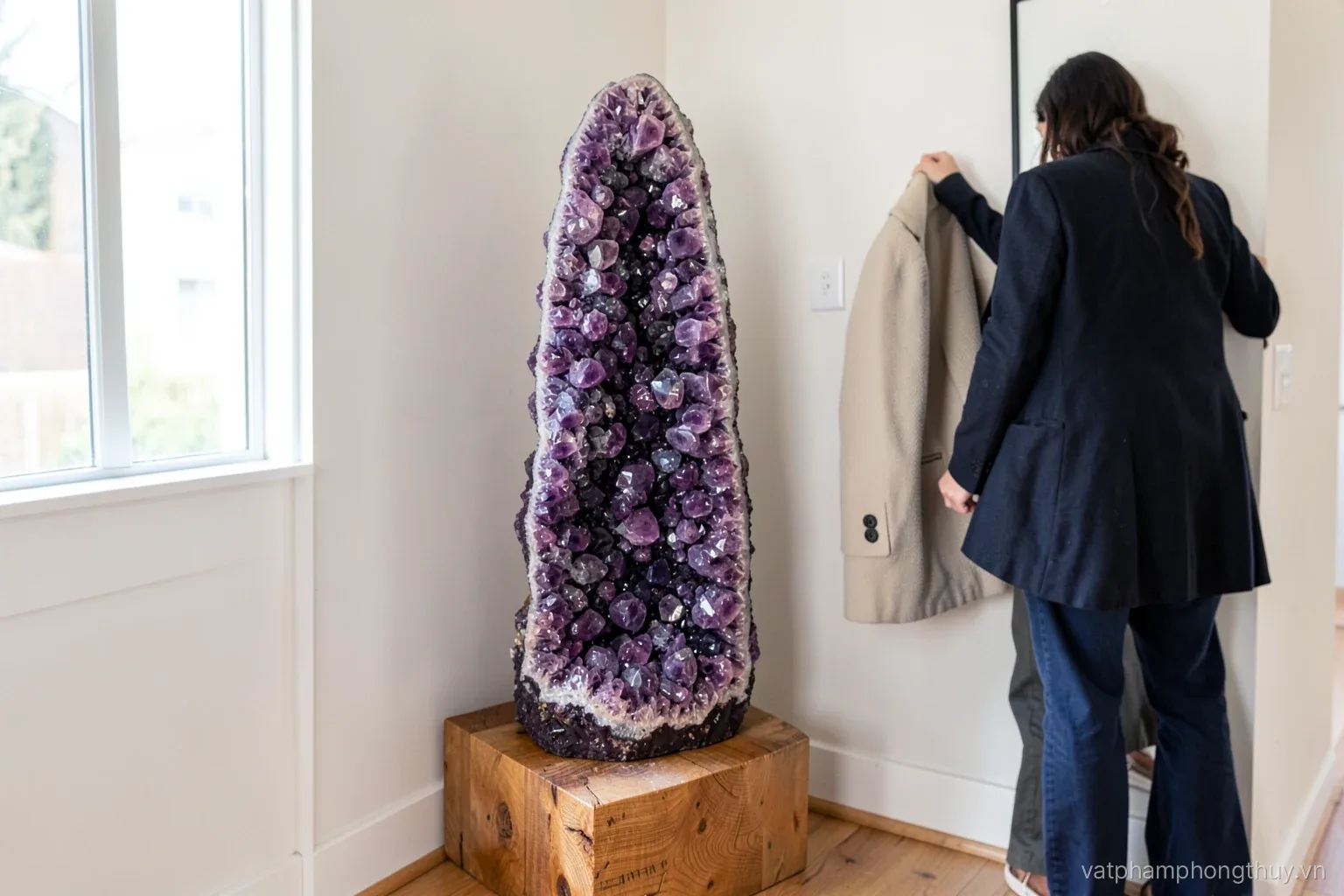 A tall amethyst geode standing on a low wooden plinth in the corner of a bright apartment entryway, a person seen from behind hanging a coat