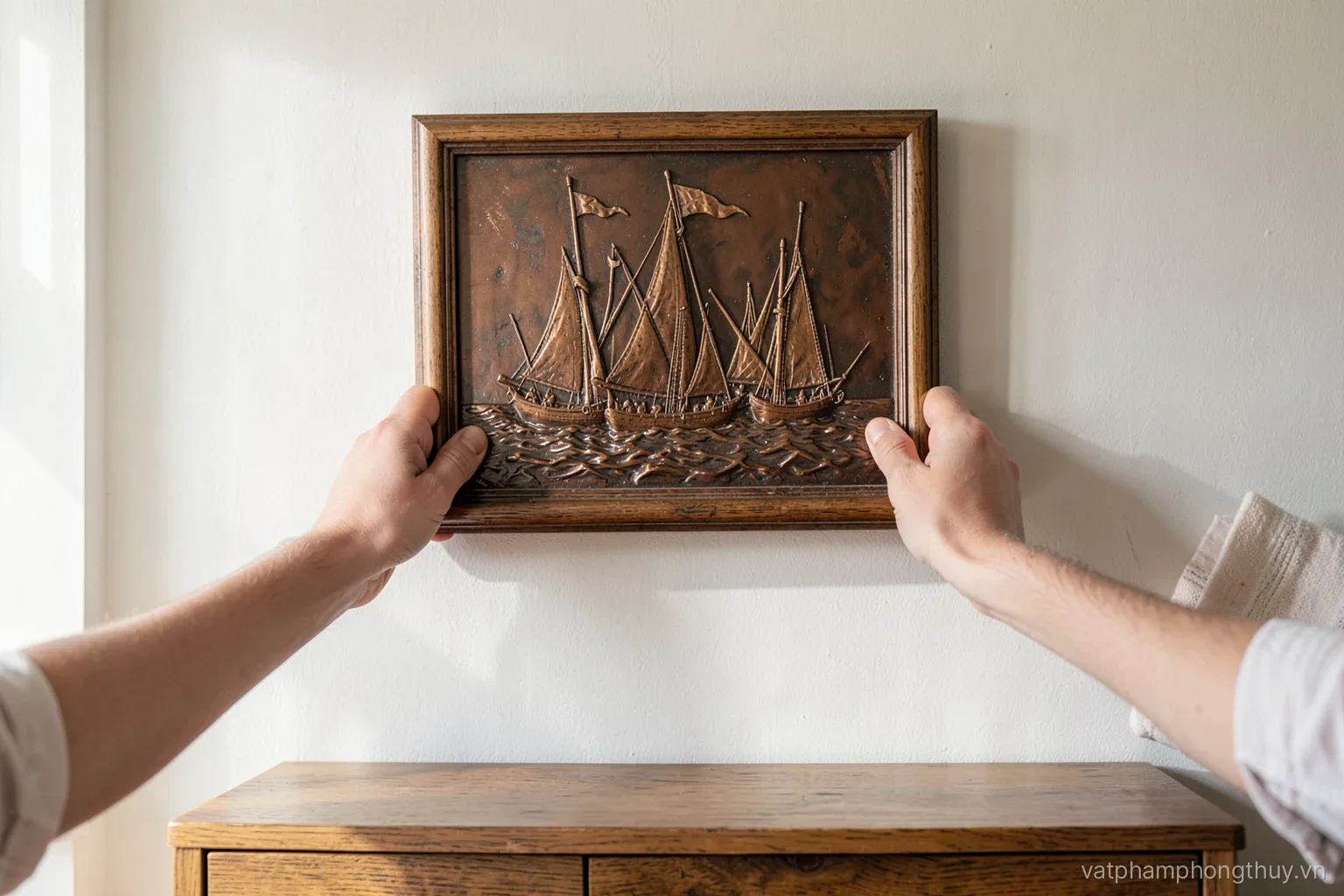 Side view of two hands leveling a framed copper relief of sailing boats on a pale living room wall above a low wooden sideboard, afternoon light