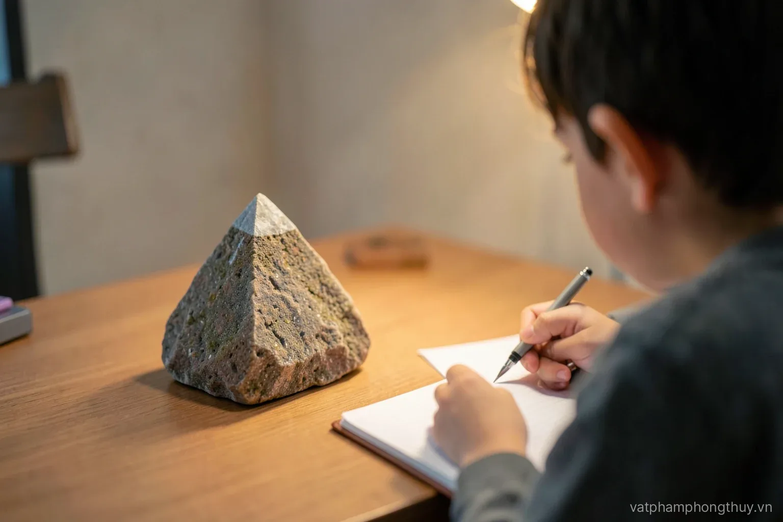 A slim hexagonal quartz point standing beside a small stone pyramid on a study desk, a child seen from behind writing in a notebook, soft lamp light