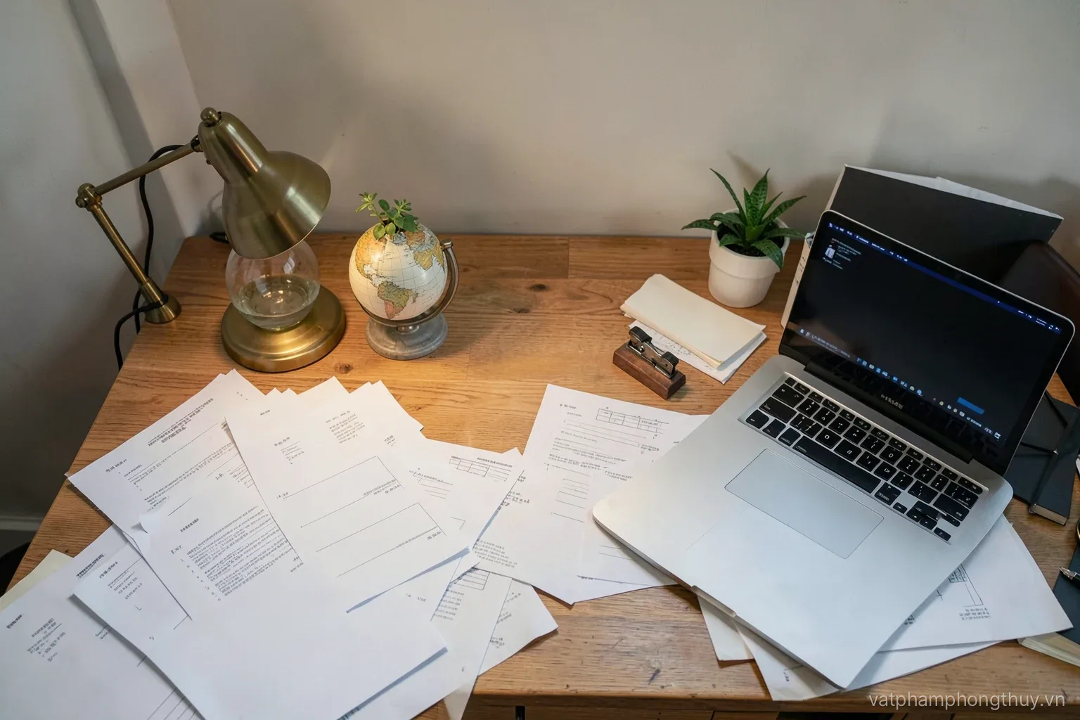 Overhead view of a cluttered shipping paperwork desk with a small brass globe paperweight holding blank forms, a stamp pad and a laptop, evening desk lamp