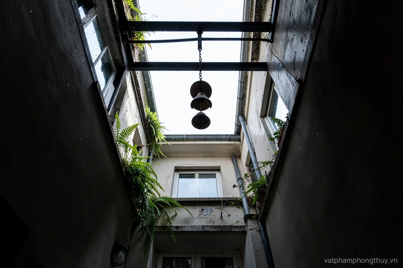 Looking straight up a narrow townhouse light well, a bronze wind chime hanging from a steel beam, ferns on ledges, rain-darkened walls and pale sky above