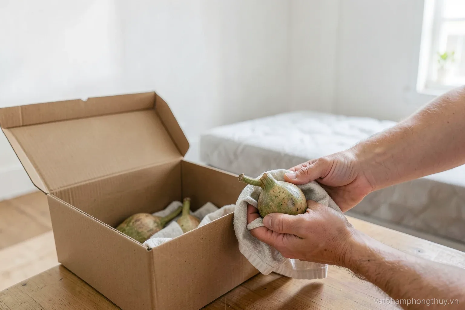 Hands wrapping a small stone gourd in a soft cloth beside an open moving box in a bright rented room, bare white walls and a folded mattress behind