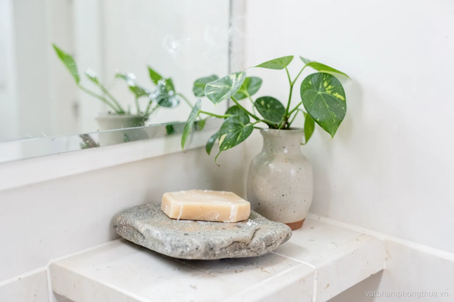 Bright small bathroom corner, a smooth river stone tray holding a bar of soap beside a ceramic vase with a green pothos on a tiled shelf, steam on the mirror
