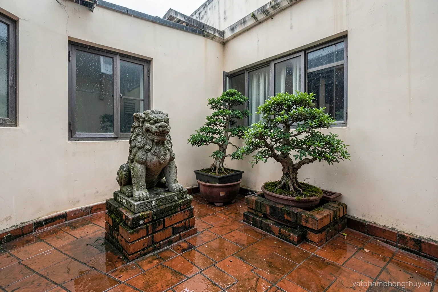 Wide view of a small Vietnamese front yard after rain, a weathered granite lion on a low brick plinth beside potted bonsai, wet tiles reflecting soft overcast light